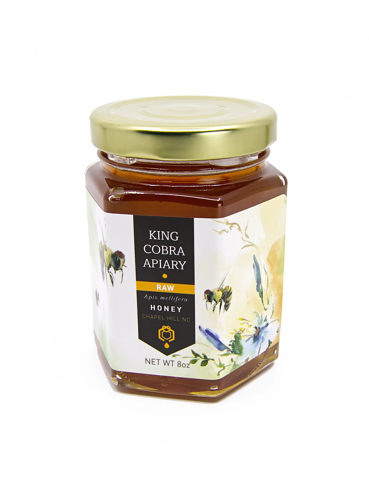 A hexagonal glass jar of King Cobra Apiary Raw Wildflower Honey with a gold metal lid and a cream-colored label featuring bee and floral illustrations, set against a plain white background.