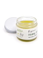 An open glass jar of organic healing salve with its white lid resting beside it on a clean white background. The salve inside is a pale yellow-green, and the label reads 'Organic Healing Salve' by 'Oil of Joy Apothecary'.